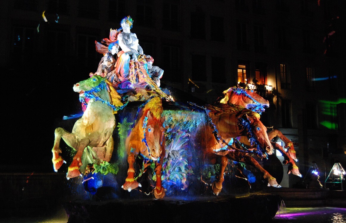 Il. 2. Fotografie przedstawiające fontannę autorstwa Frédérica Auguste’a Bartholdiego stojącą na Placu Terreaux w Lyonie. Z lewej strony – widok fontanny za dnia; z prawej strony – jedna z odsłon fontanny w porze nocnej w trakcie festiwalu świateł (Fête des Lumières) w roku 2010. W trakcie pokazów fontanna dynamicznie zmieniała swoją barwę. Nałożenie kolorów na poszczególne elementy fontanny, odmieniające jej jednolity, monumentalny wygląd, nie jest fizyczną ingerencją w tkankę obiektu, a efektem zrzutowania na niego barwnego światła (zdjęcie nocne 2010, K. Wlazło-Malinowska, zdjęcie dzienne – źródło: https://upload.wikimedia.org/wikipedia/commons/5/5e/Fontaine_Bartholdi_Lyon_16.jpg).